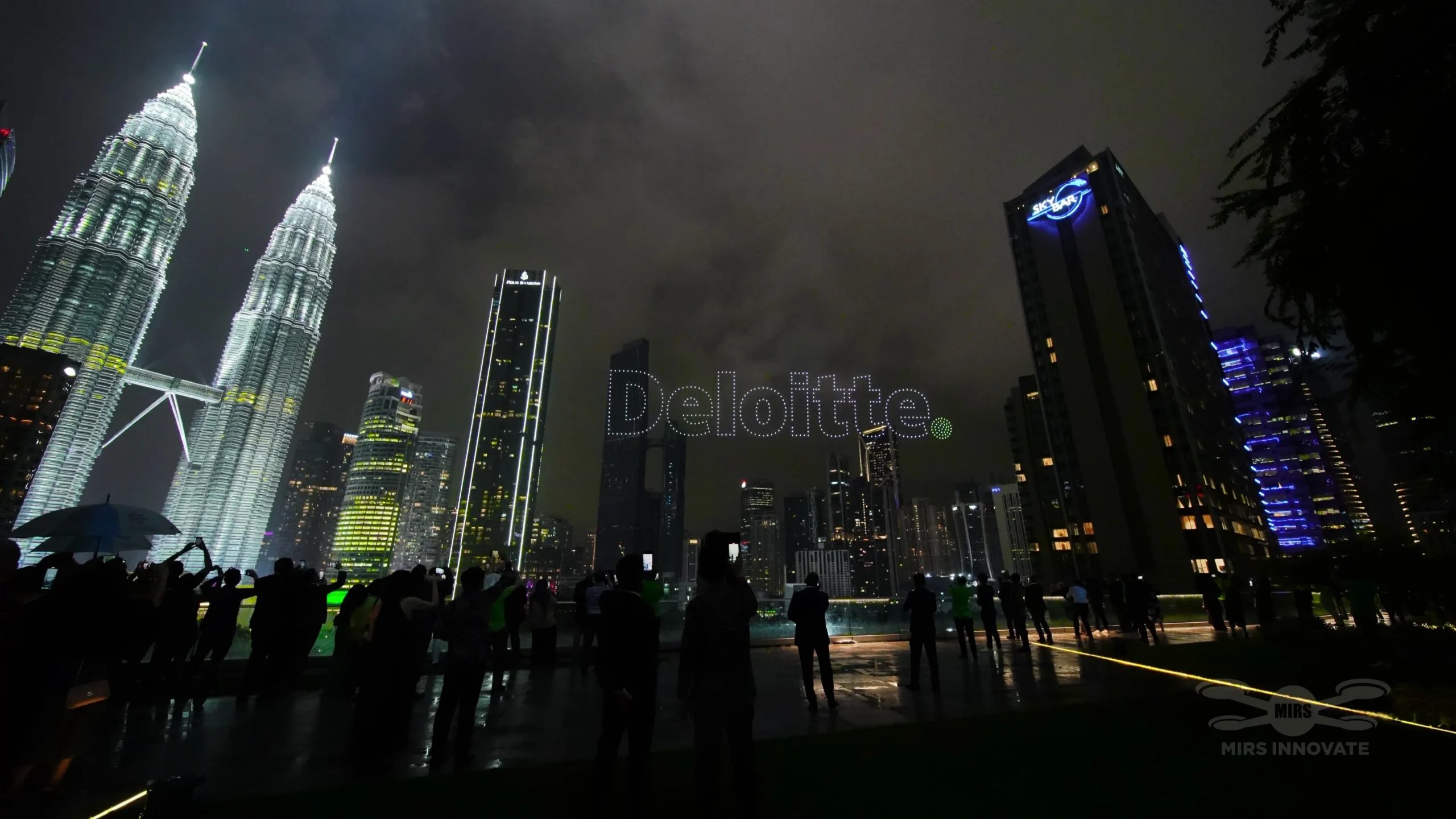 Deloitte drone text formation above Petronas Twin Towers with crowd below KLCC Park